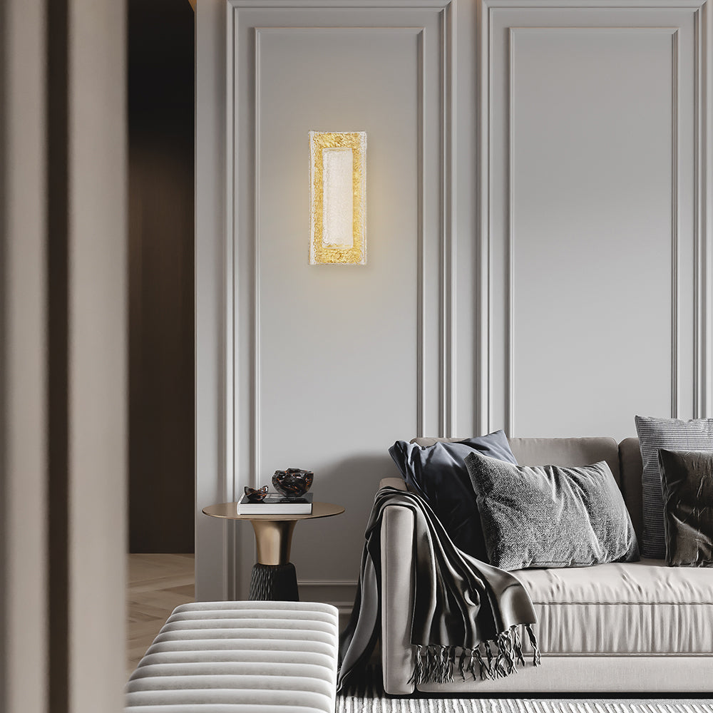 The Zeamas Wall Sconce's translucent ice-like glass surface casts a soft, ambient light in your living room, creating a serene atmosphere that complements the space's design with its aged brass backplate and energy-efficient LED lighting.